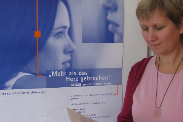 Eine Frau liest ein Dokument. Hinter ihr hängt ein Plakat an der Wand mit der Aufschrift „Mehr als das Herz gebrochen. Gewalt macht Frauen krank“.